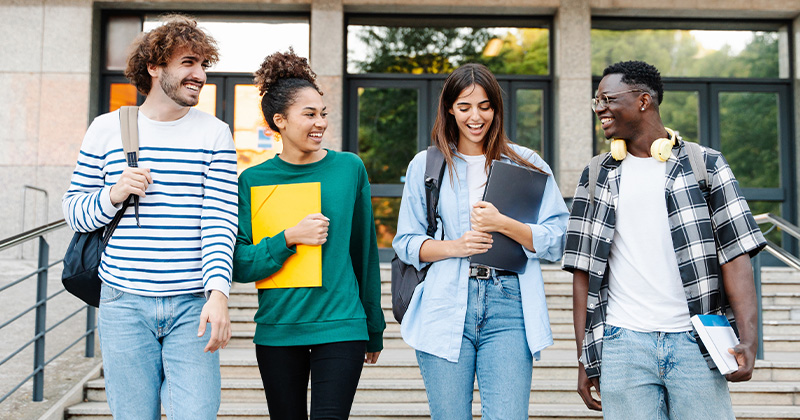 four diverse students walking together outside a school building engaged in conversation smiling and holding books this group represents a lively college community exploring academic opportunities 3 friends studying together